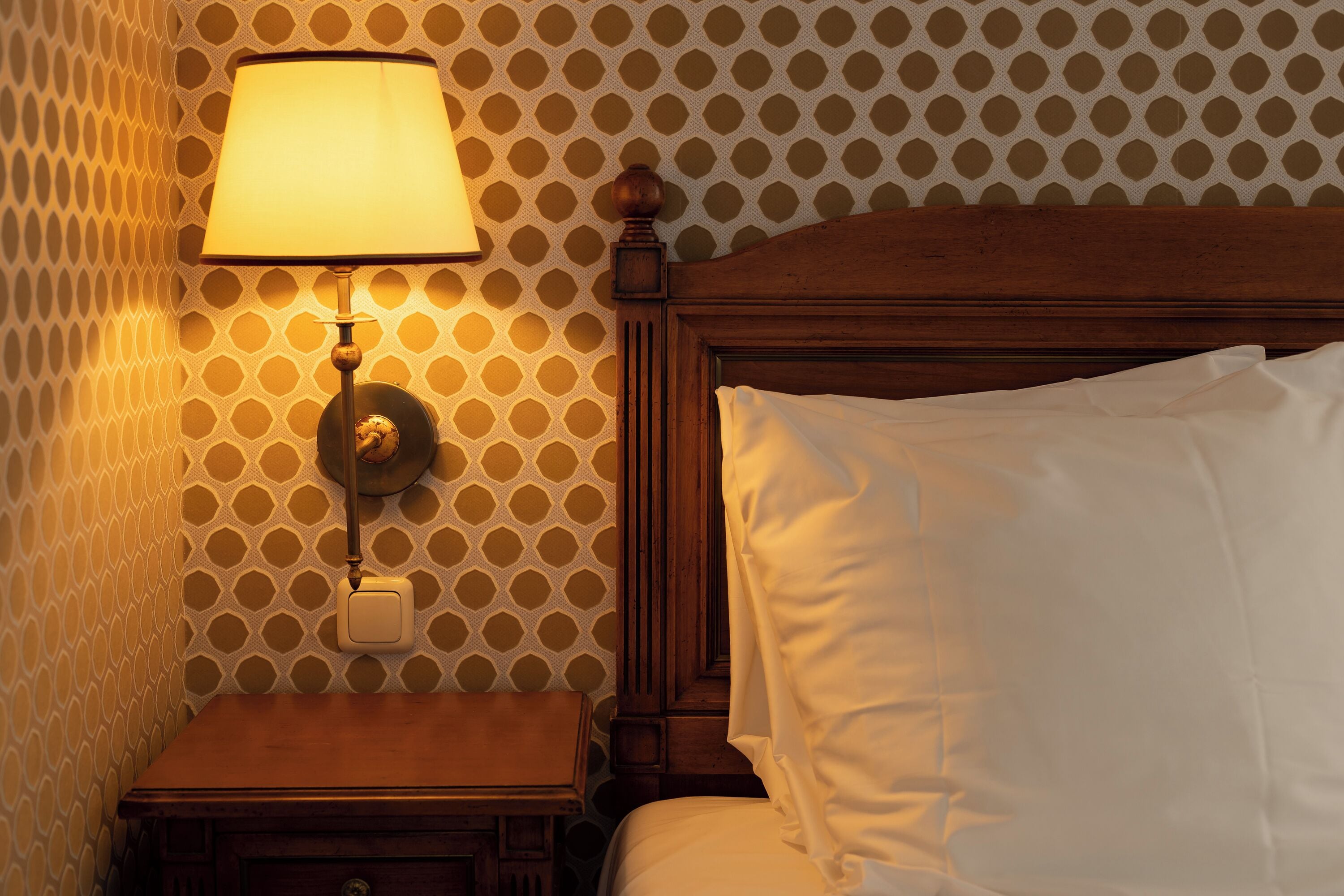 A neatly made bed with white linens, a wooden headboard, and a wall-mounted lamp.