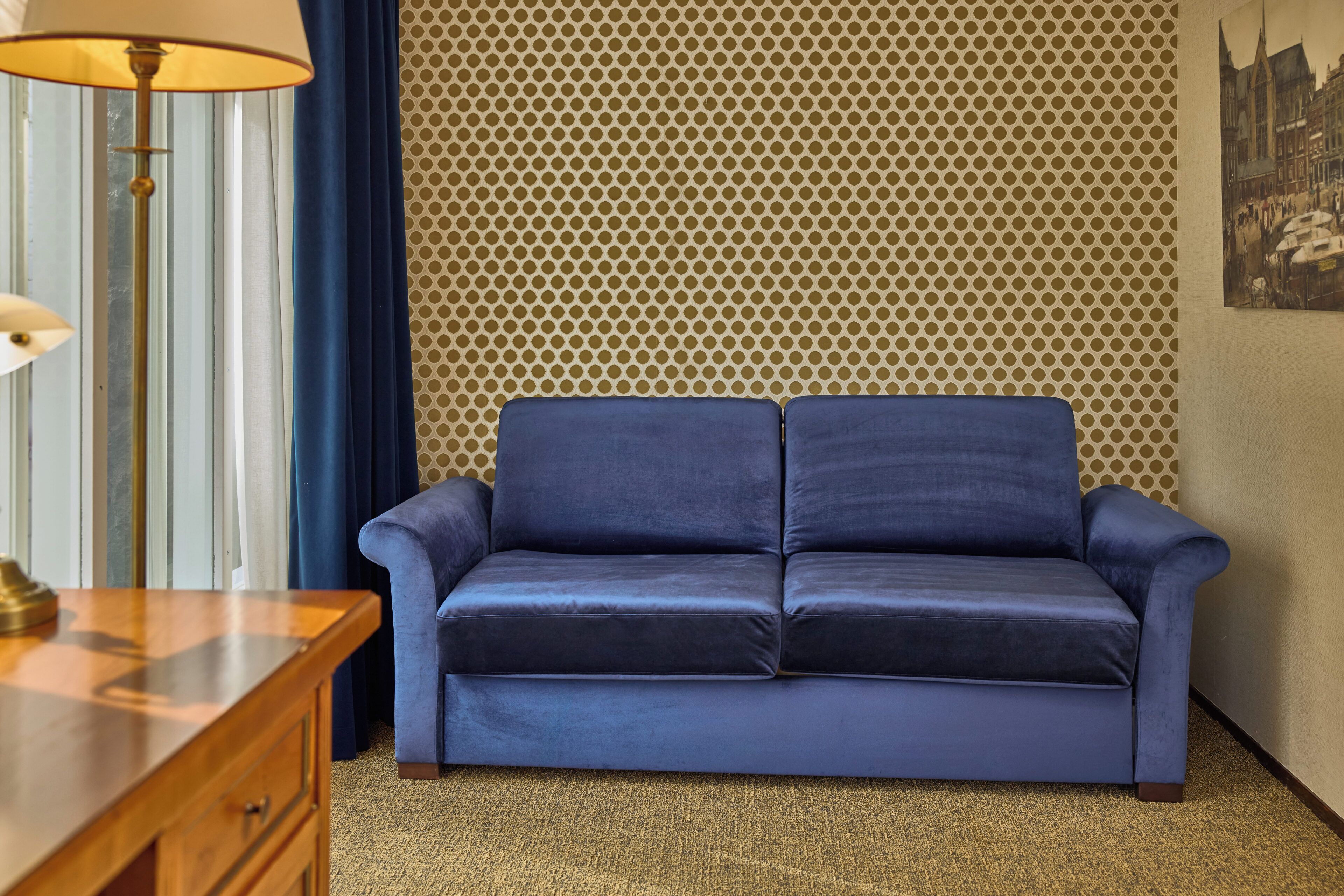 A room with a blue sofa, a wooden side table, a patterned wall, and a lamp.