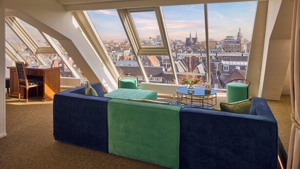 Une pièce aux hauts plafonds offrant une vue sur la ville, avec une table ronde et des sièges aux couleurs verte et bleue.