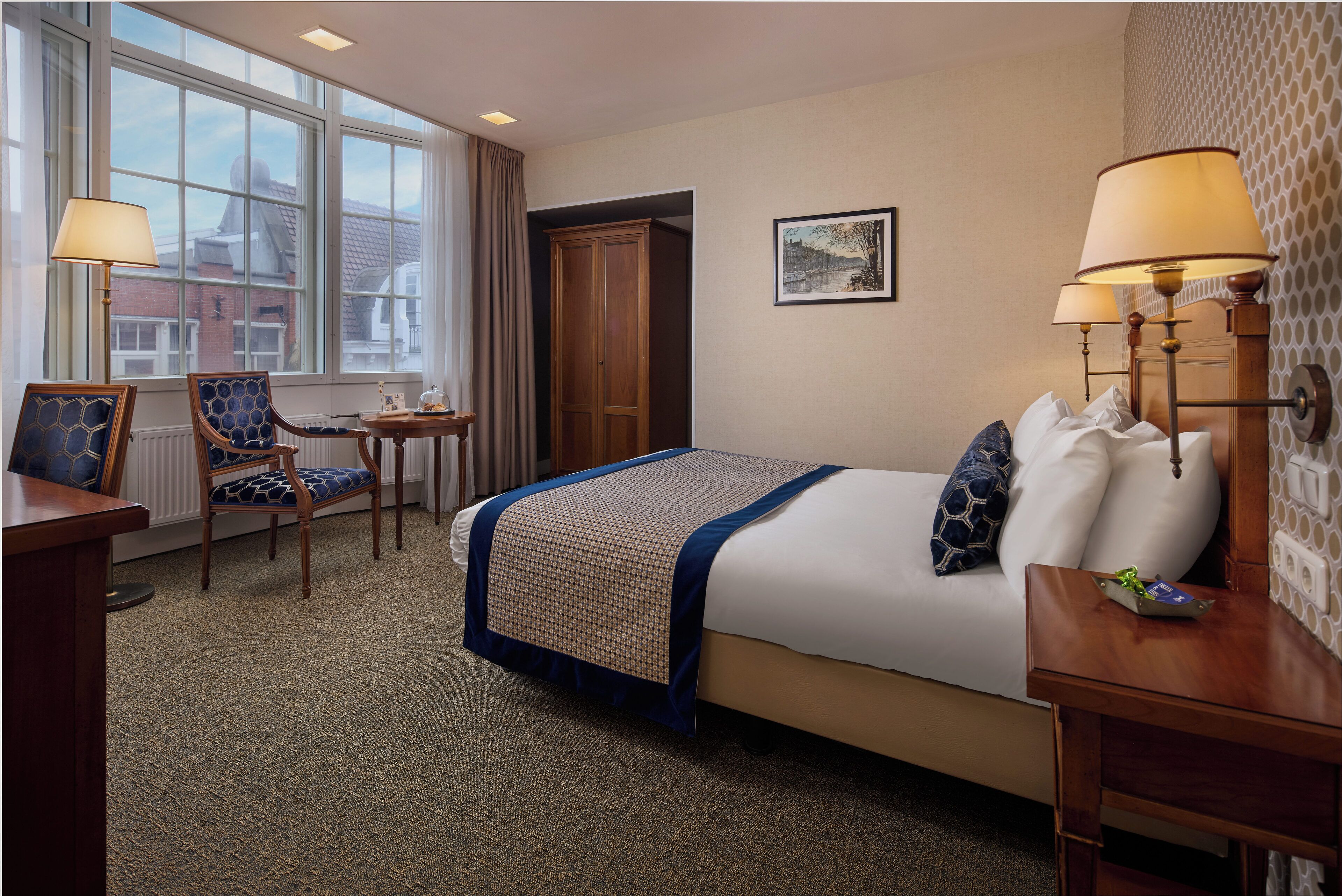 A hotel room with a bed, a desk, a chair, a window with a view, and a lamp.
