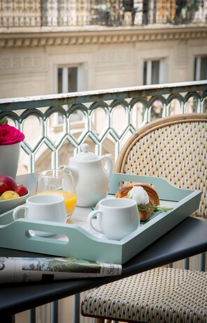 Un plateau de petit-déjeuner sur un balcon, avec une chaise en osier, un pichet blanc, un verre de jus d’orange et un croissant.