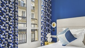 A bedroom with a bed, curtains, a bedside table, and a view of a building through a window. - Hôtel Elysées Opéra (Paris)