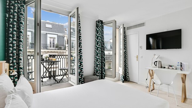 A hotel room with a bed, a desk, a TV, and a balcony with a view of buildings. - Hôtel Elysées Opéra (Paris)