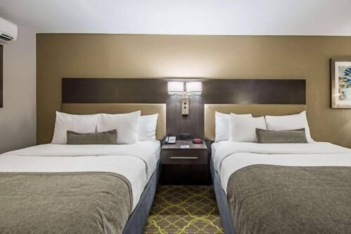 A hotel room with two beds, a nightstand with a phone and a lamp, and a wall-mounted light fixture.