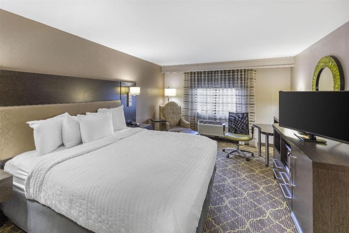 A hotel room with a large bed, a desk with a flat-screen TV, a chair, and a window with curtains.