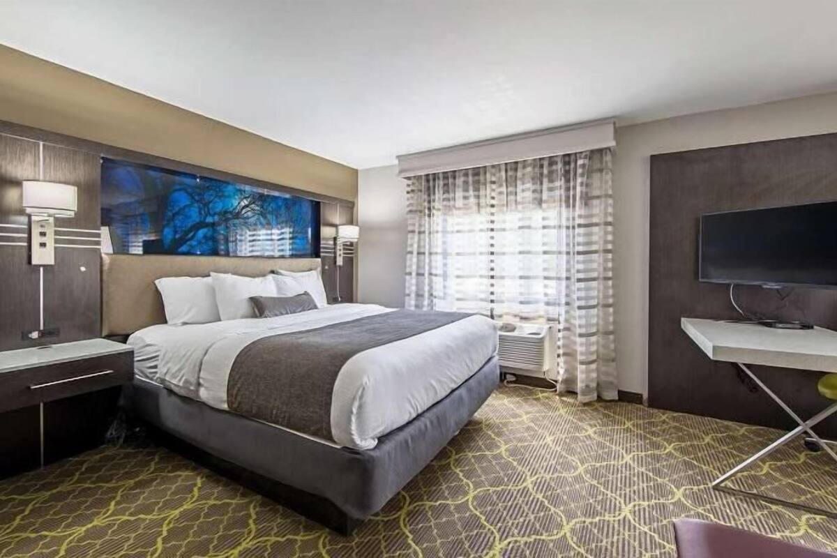 A hotel room with a bed, bedside tables, a desk with a chair, a television, and a large window with curtains.