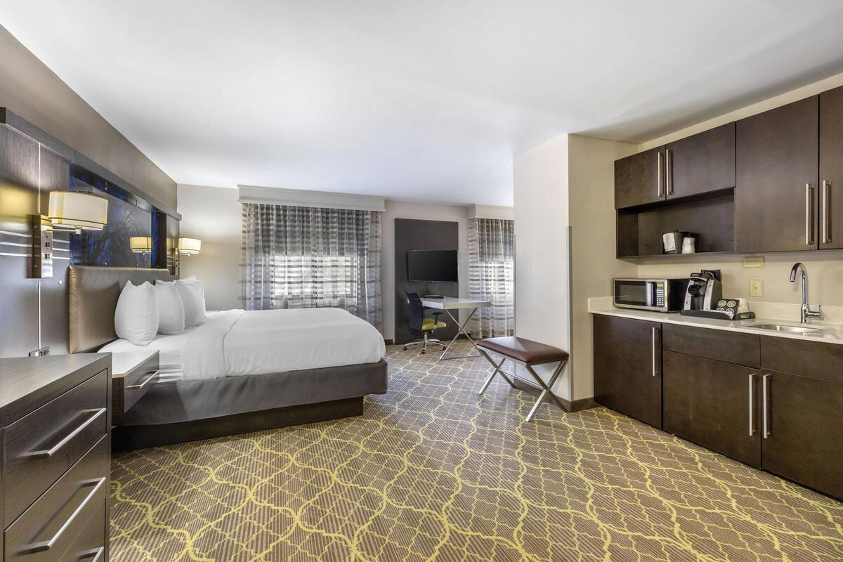 A hotel room with a bed, desk, kitchenette, and a patterned carpet.