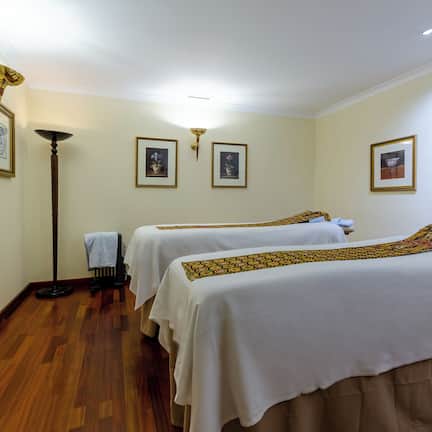 A hotel room with two beds, a standing lamp, framed pictures on the wall, and a wooden floor.