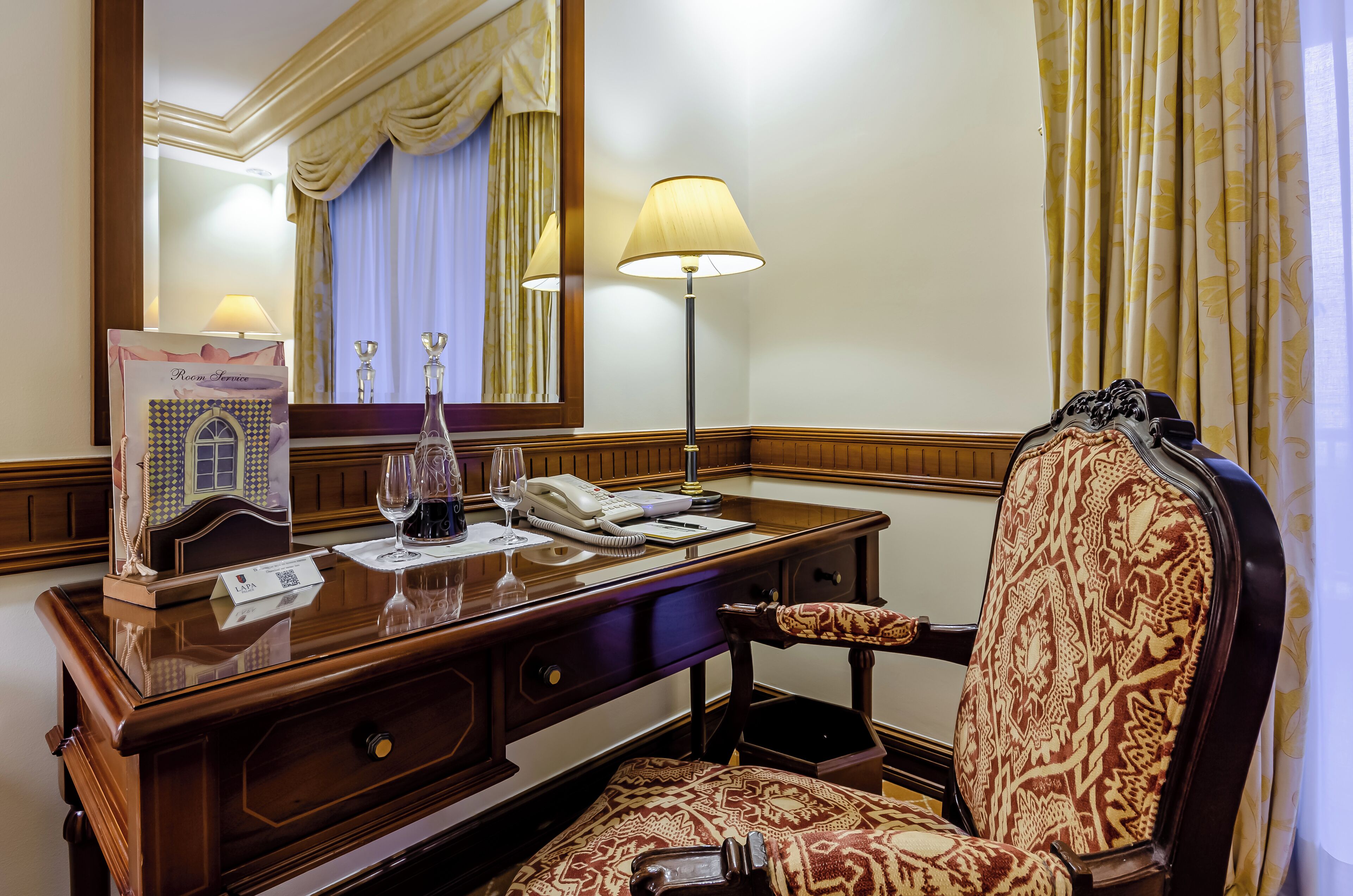Habitación de hotel con un escritorio de madera, un teléfono, un vaso y un folleto.