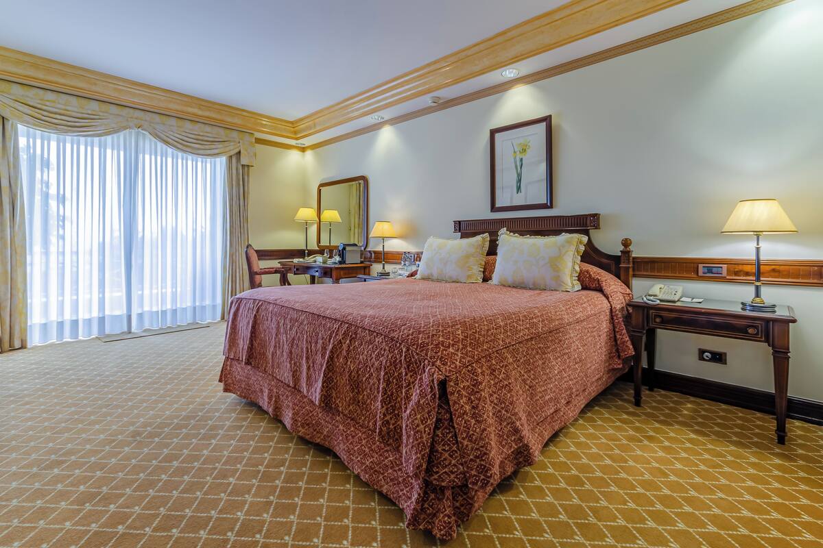 A hotel room with a large bed, two bedside tables, a nightstand, a desk, a chair, a window with curtains, and a framed picture on the wall.