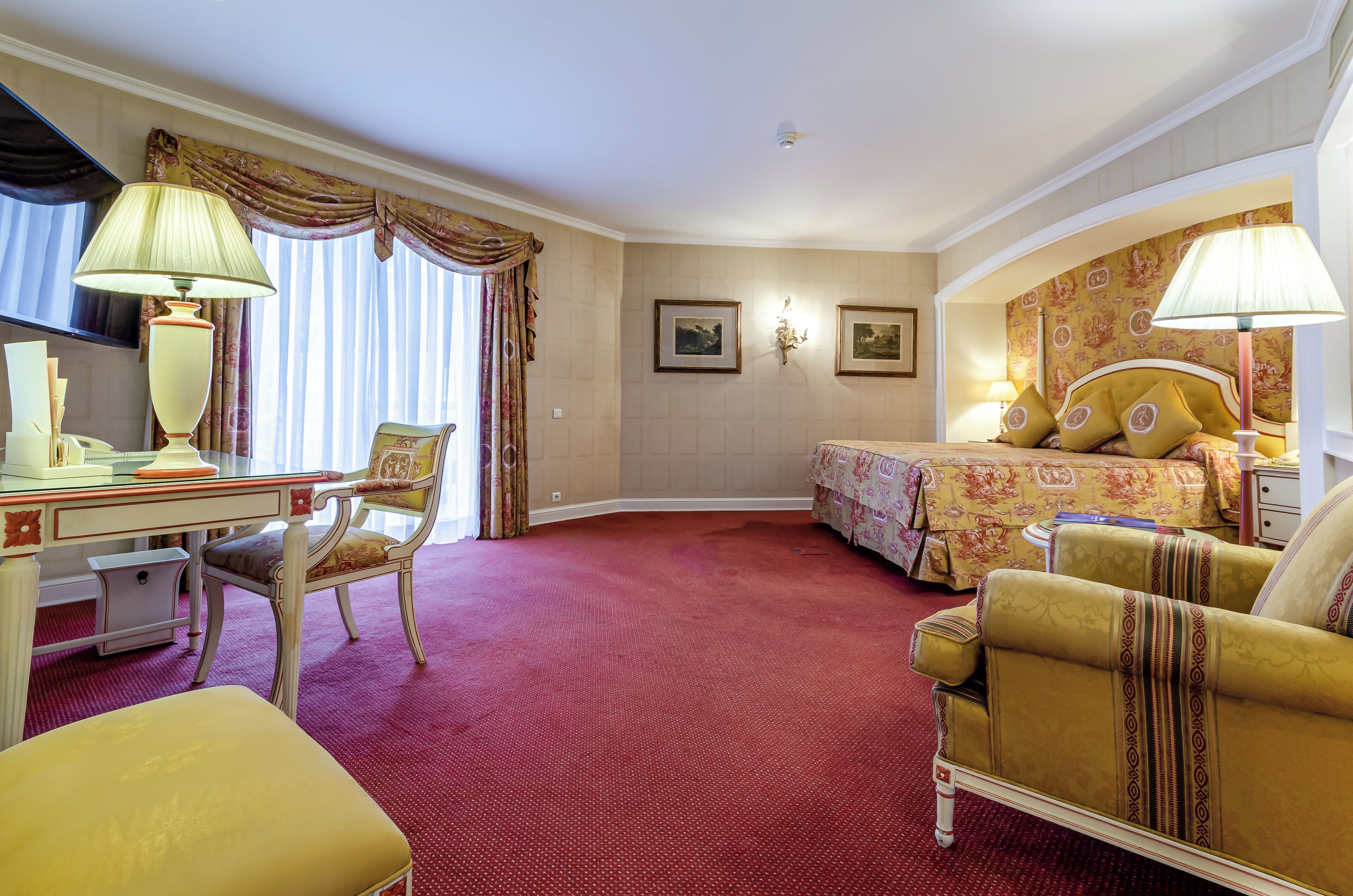 Une chambre d’hôtel équipée d’un bureau, de deux chaises, d’un lit avec une couvre-lit à motifs floraux et d’une moquette rouge.