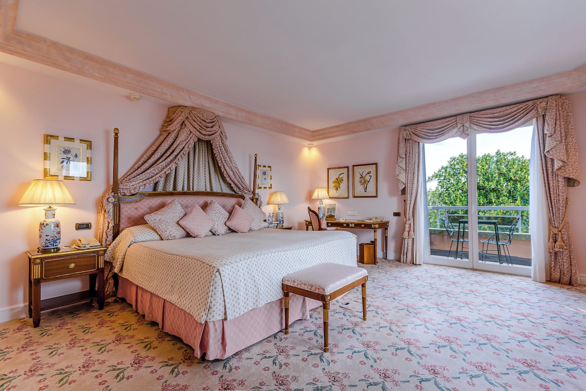 A bedroom with a four-poster bed, a bench, a desk, and a balcony with a view.
