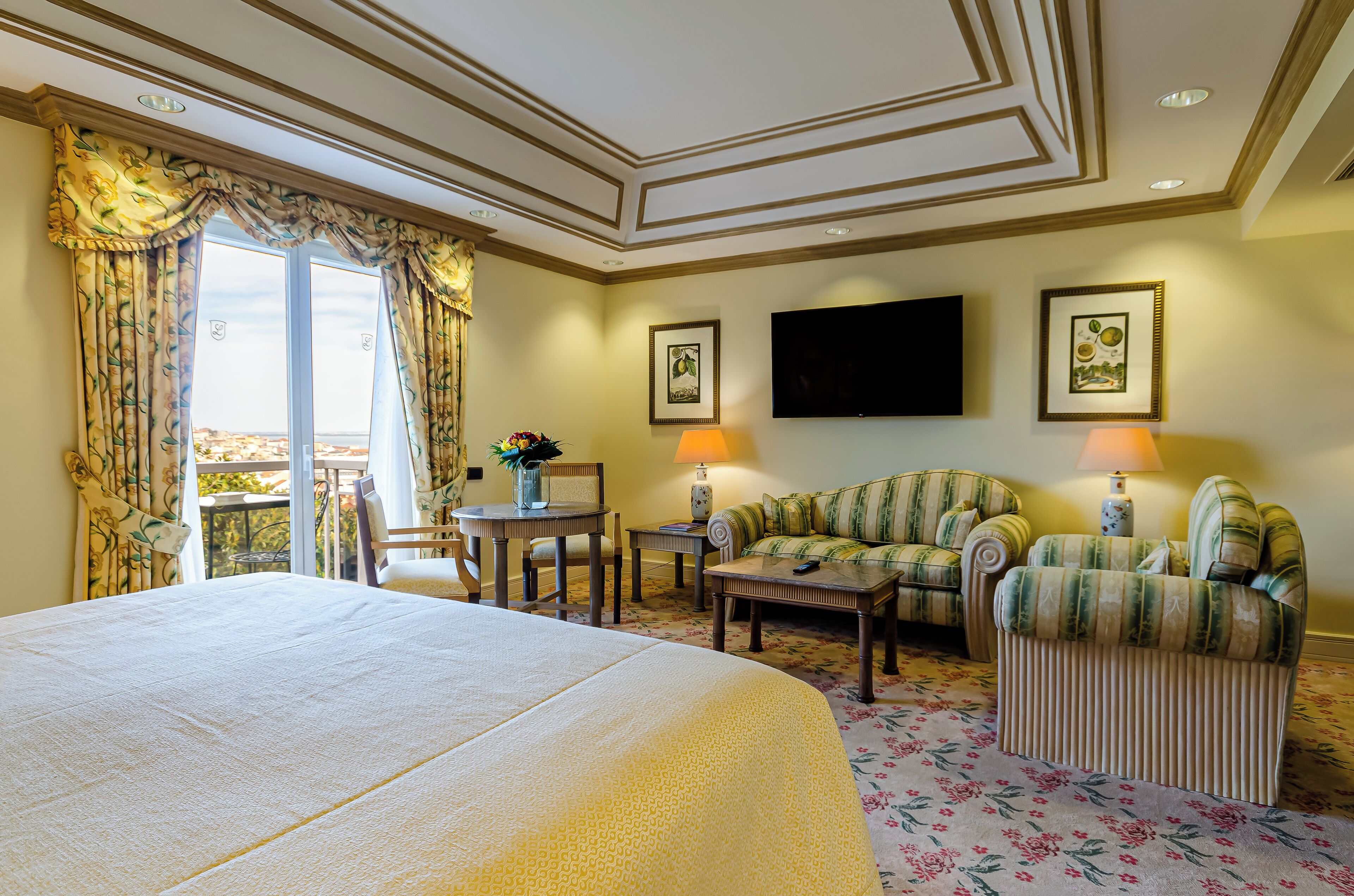 A hotel room with a large bed, two armchairs, a sofa, a small table, a TV, and a balcony with curtains.