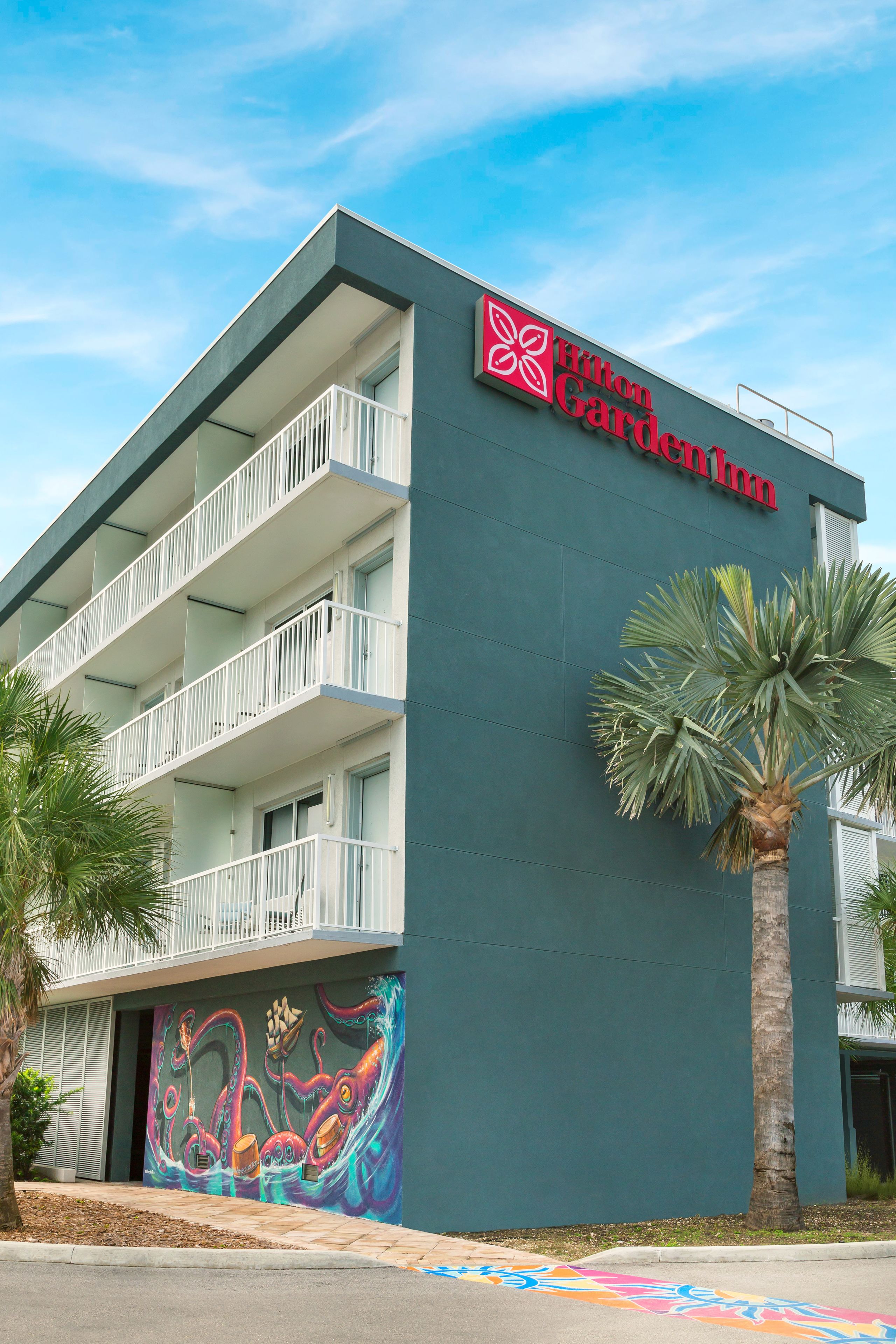 A Hilton Garden Inn building with a mural of an octopus on the side, balconies, and palm trees.