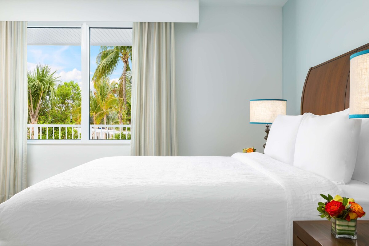 A hotel room with a bed, a nightstand, a lamp, a window with curtains, and a view of palm trees.
