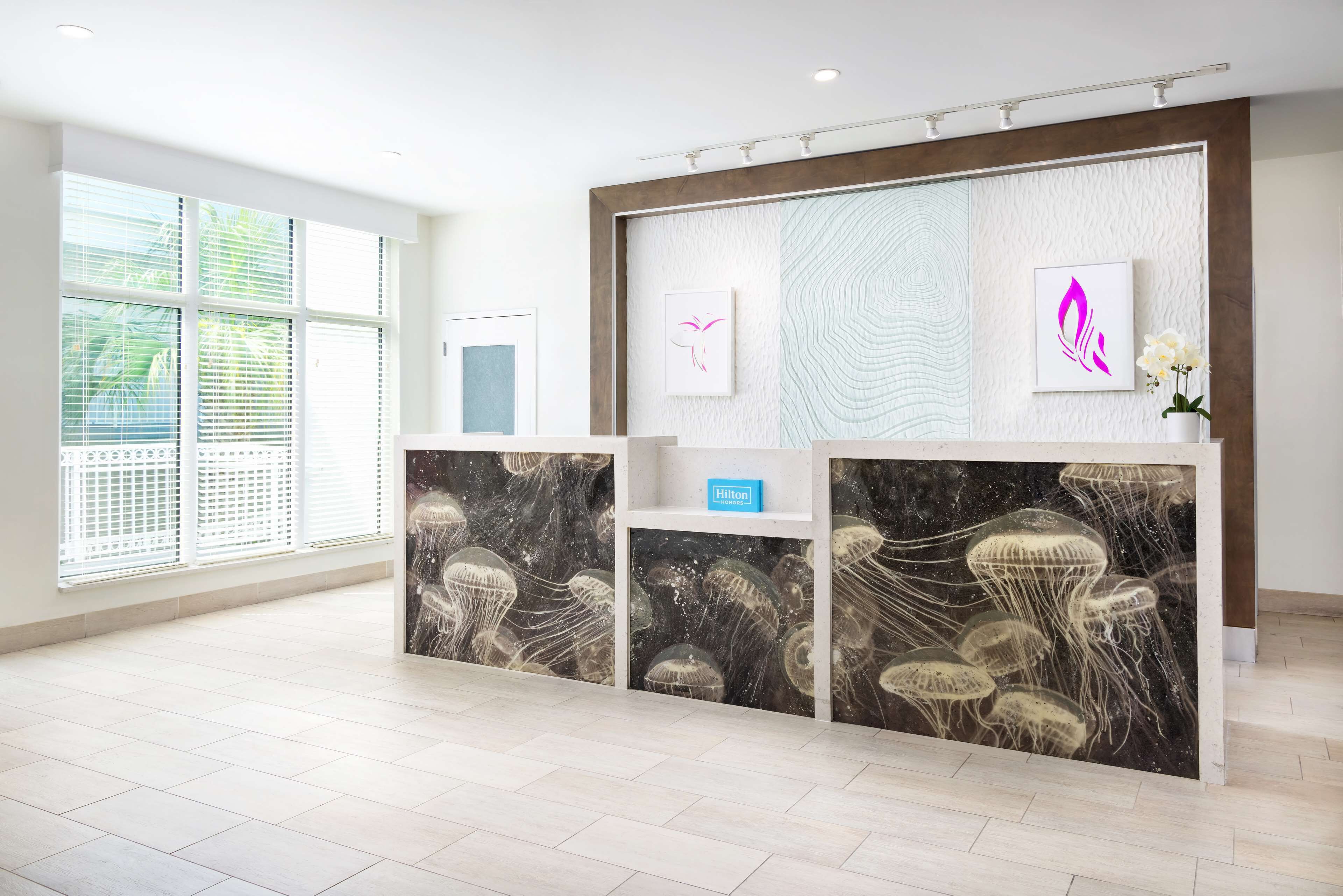 A hotel reception area with a modern design, featuring a marble reception desk, a large window with blinds, and artwork on the walls.