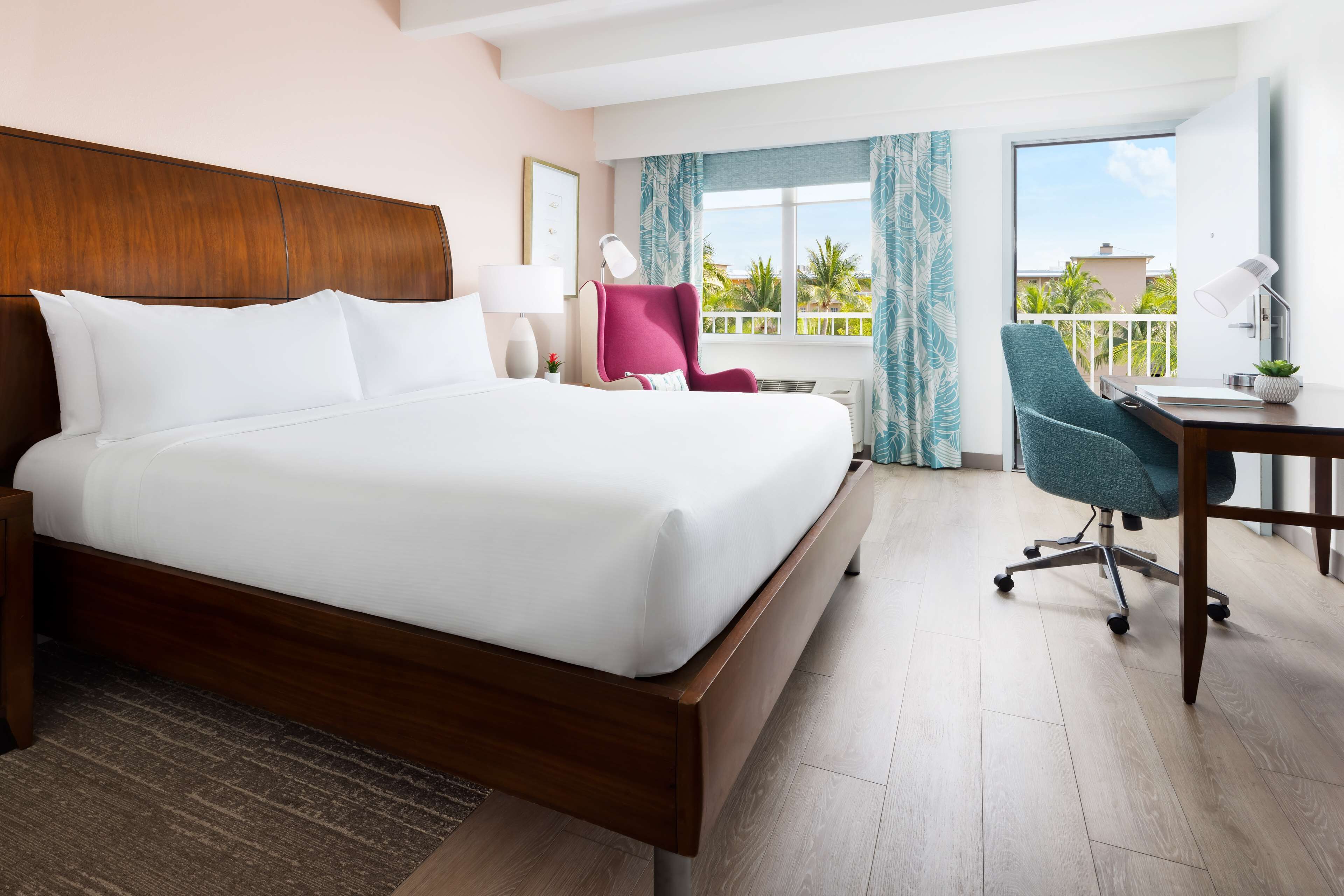 A hotel room with a large bed, a desk with a chair, a window with curtains, and a view of palm trees.