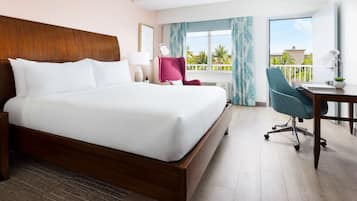 A hotel room with a large bed, a desk with a chair, a window with curtains, and a view of palm trees.