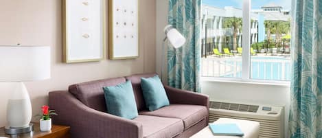A hotel room with a bed, a sofa, a side table with a lamp, a window with a view of a pool and palm trees, and a ceiling fan.