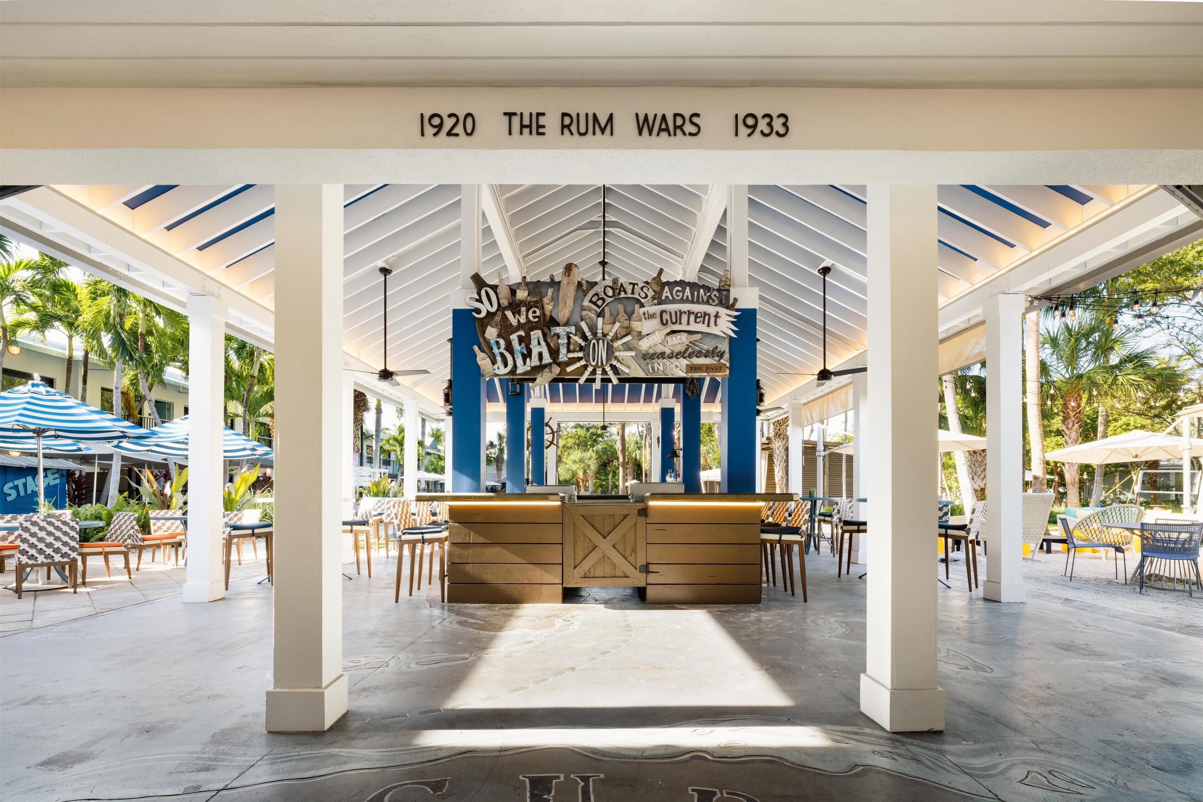 Un espace extérieur abrité doté d’un bar central en bois, de palmiers et d’un plafond sur lequel est affiché un panneau indiquant : « 1920, L’ÈRE DE LA CONTREBANDE DE RHUM, 1933 ».