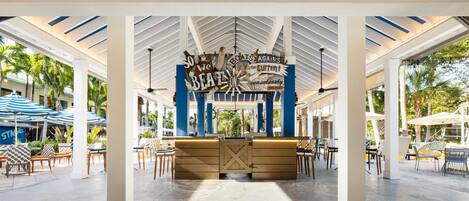 Un espace extérieur abrité doté d’un bar central en bois, de palmiers et d’un plafond sur lequel est affiché un panneau indiquant : « 1920, L’ÈRE DE LA CONTREBANDE DE RHUM, 1933 ».