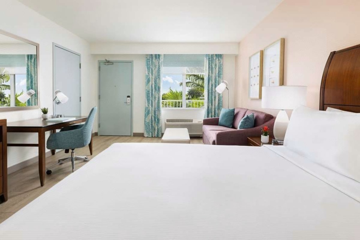 Habitación de hotel con cama, escritorio, silla, sofá, lámpara y ventana con cortinas.