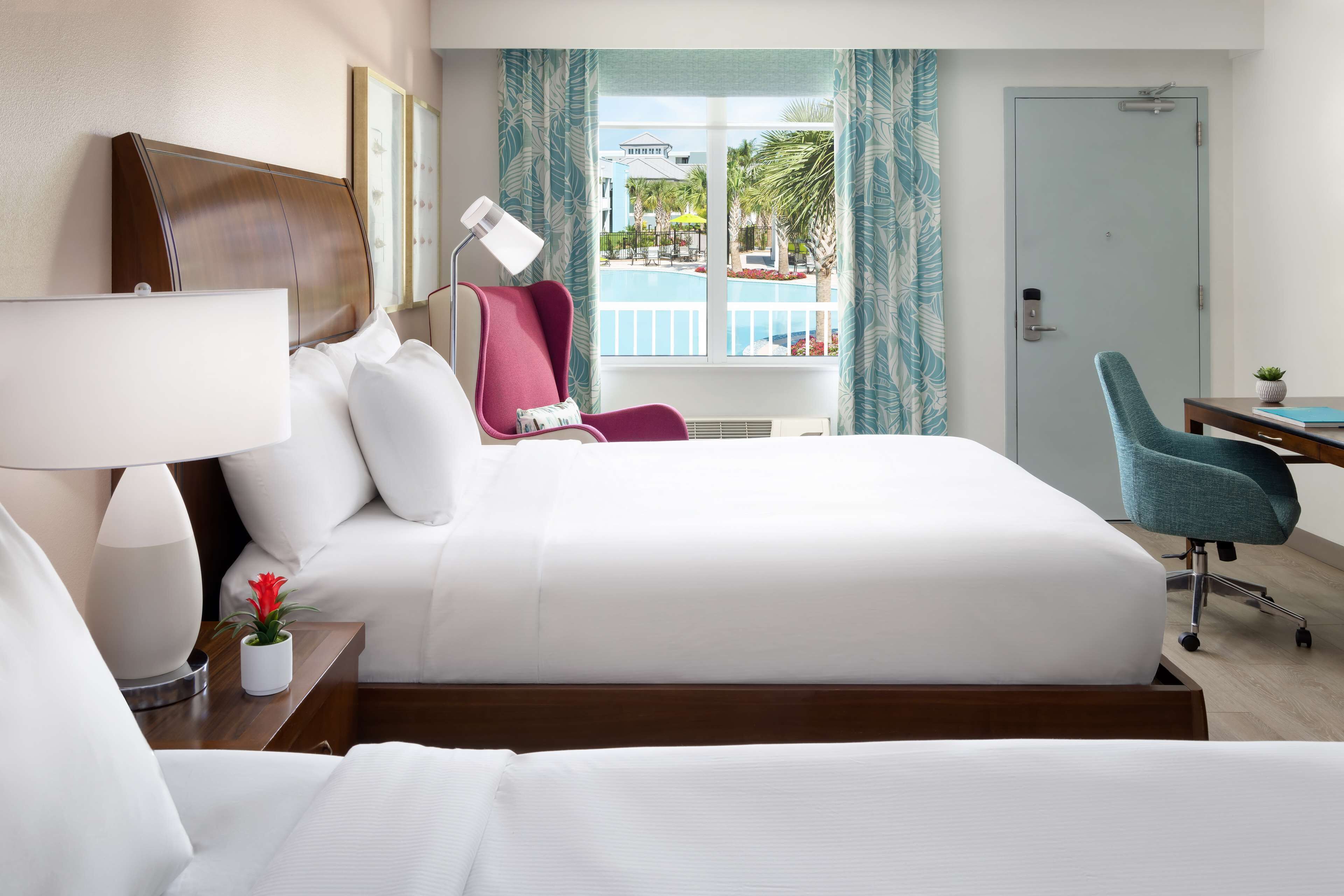 A hotel room with a large bed, a desk, a chair, a lamp, and a view of a pool and palm trees.