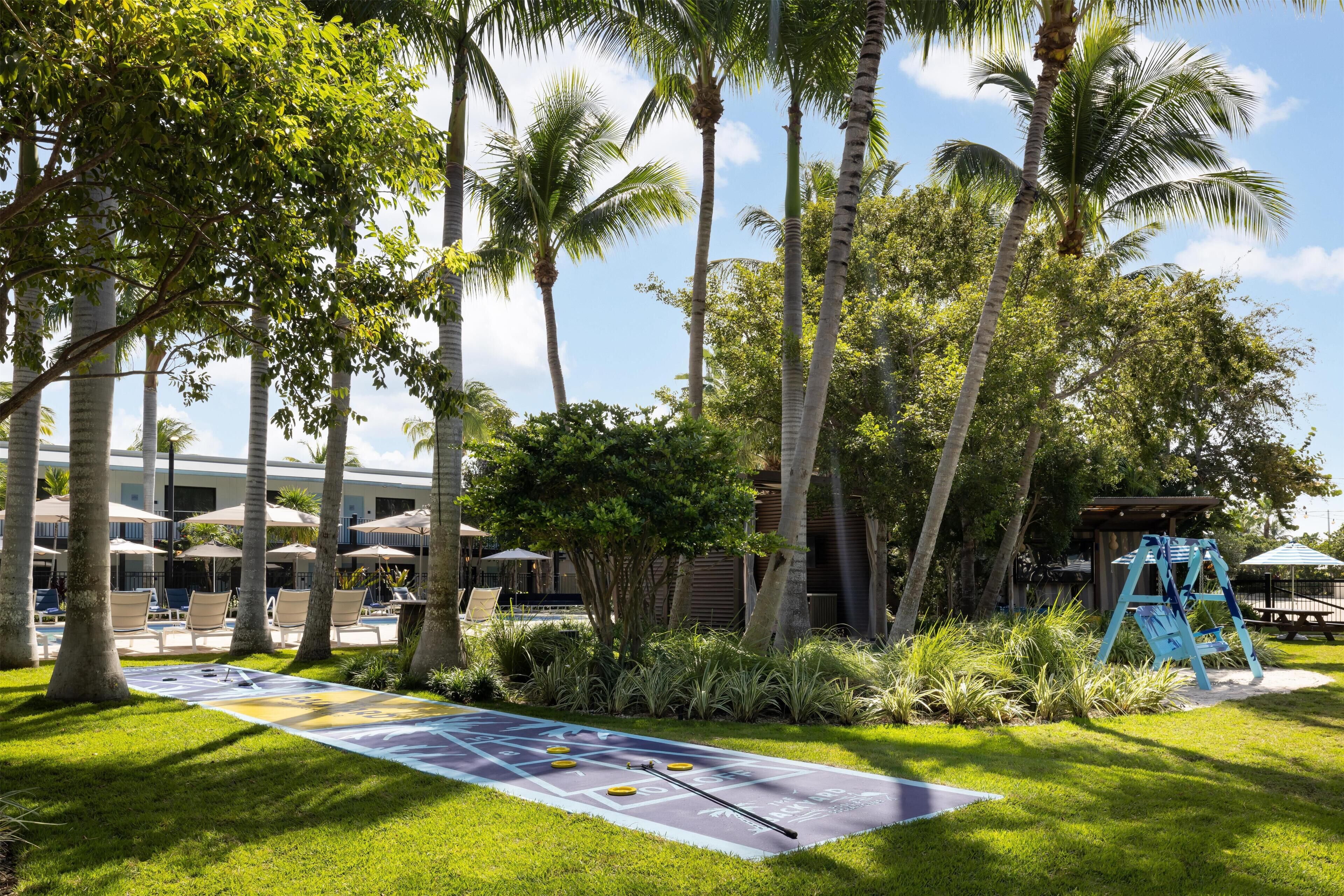 A courtyard with a shuffleboard table, lounge chairs, palm trees, and a wooden structure.