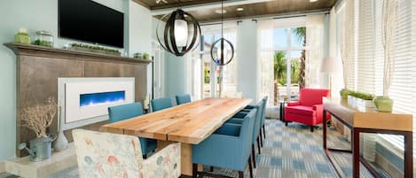 Un comedor amplio con una larga mesa de madera y sillas tapizadas en azul, un sillón rojo y una pequeña mesita con una lámpara.