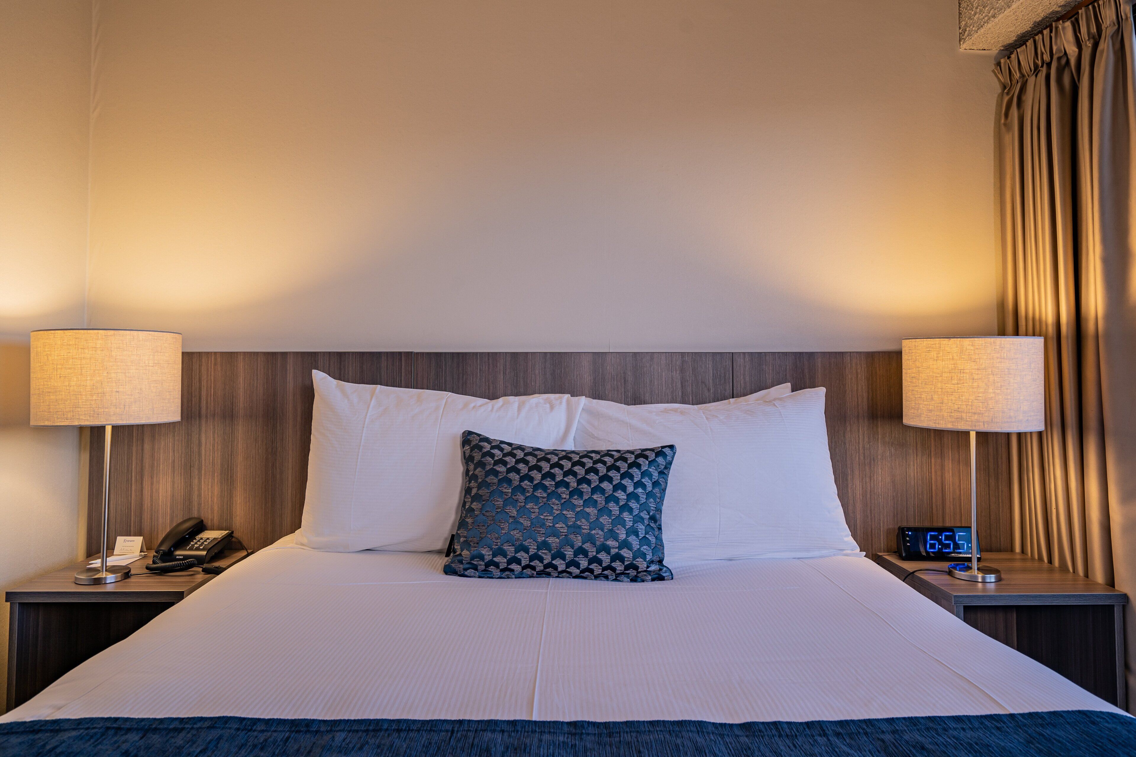 A neatly made bed with white linens, a patterned pillow, and two bedside lamps. A digital clock on the nightstand displays the time.