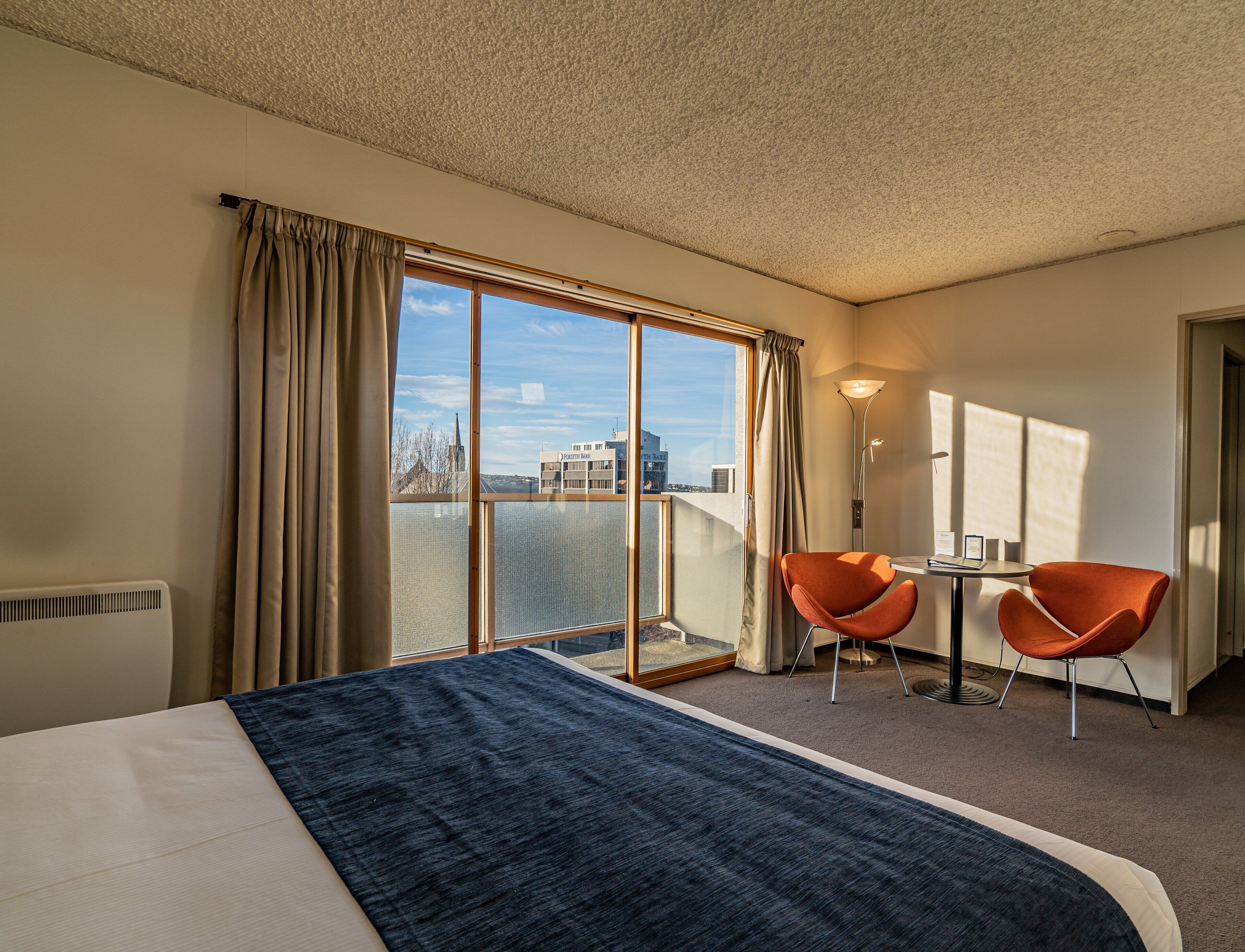 A hotel room with a large bed, two orange chairs, a small table, and a view of the city through large windows.