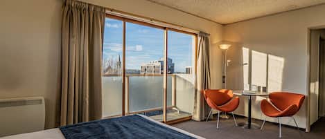 Habitación de hotel con una cama grande, dos sillones naranja, una mesita y vistas a la ciudad a través de amplios ventanales.