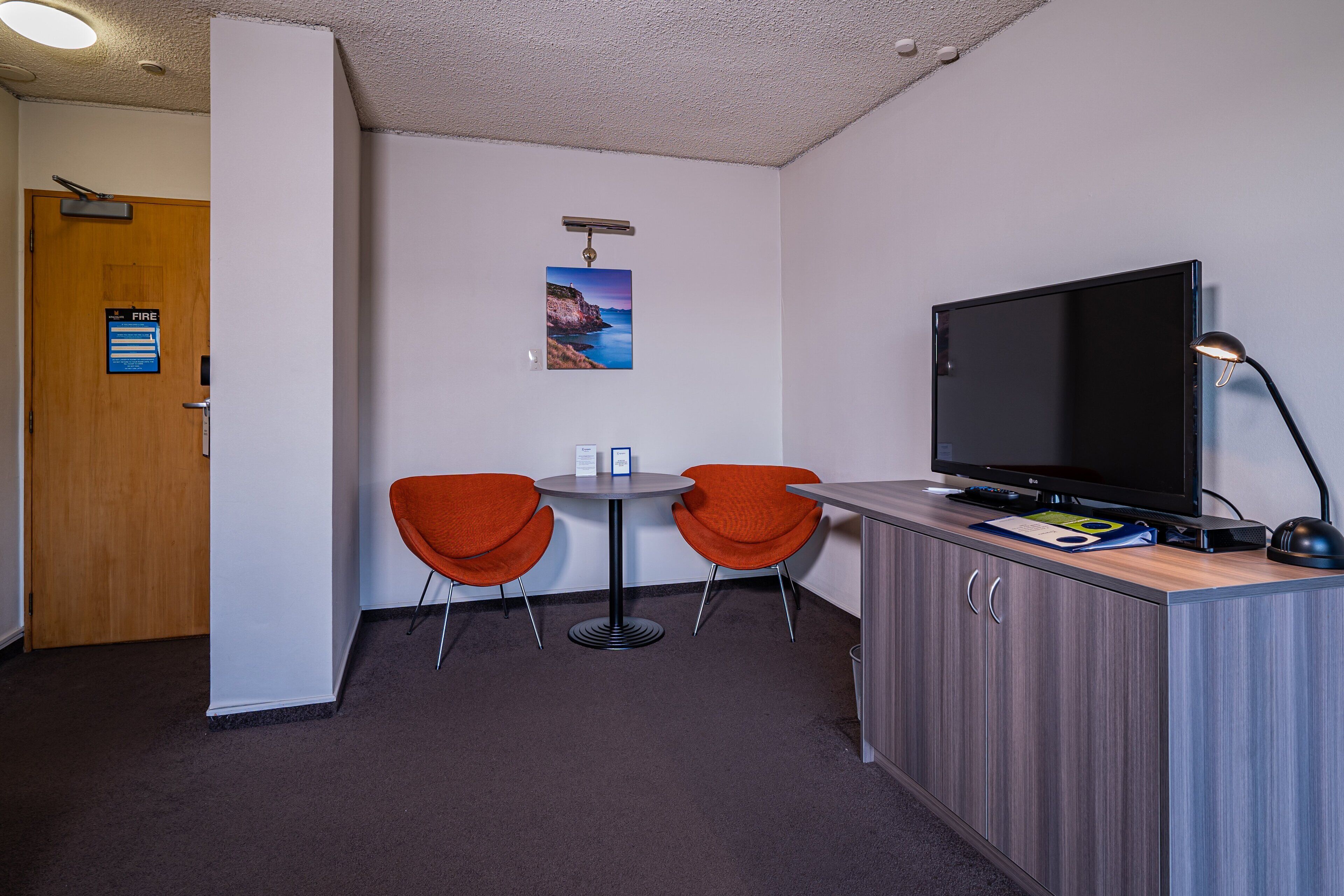Une petite pièce avec une porte en bois, un panneau indiquant la sortie de secours, deux chaises orange, une petite table, un téléviseur et un bureau avec une lampe.