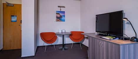 Una habitación pequeña con una puerta de madera, un letrero de salida de incendios, dos sillas color naranja, una mesita, un televisor y un escritorio con una lámpara.