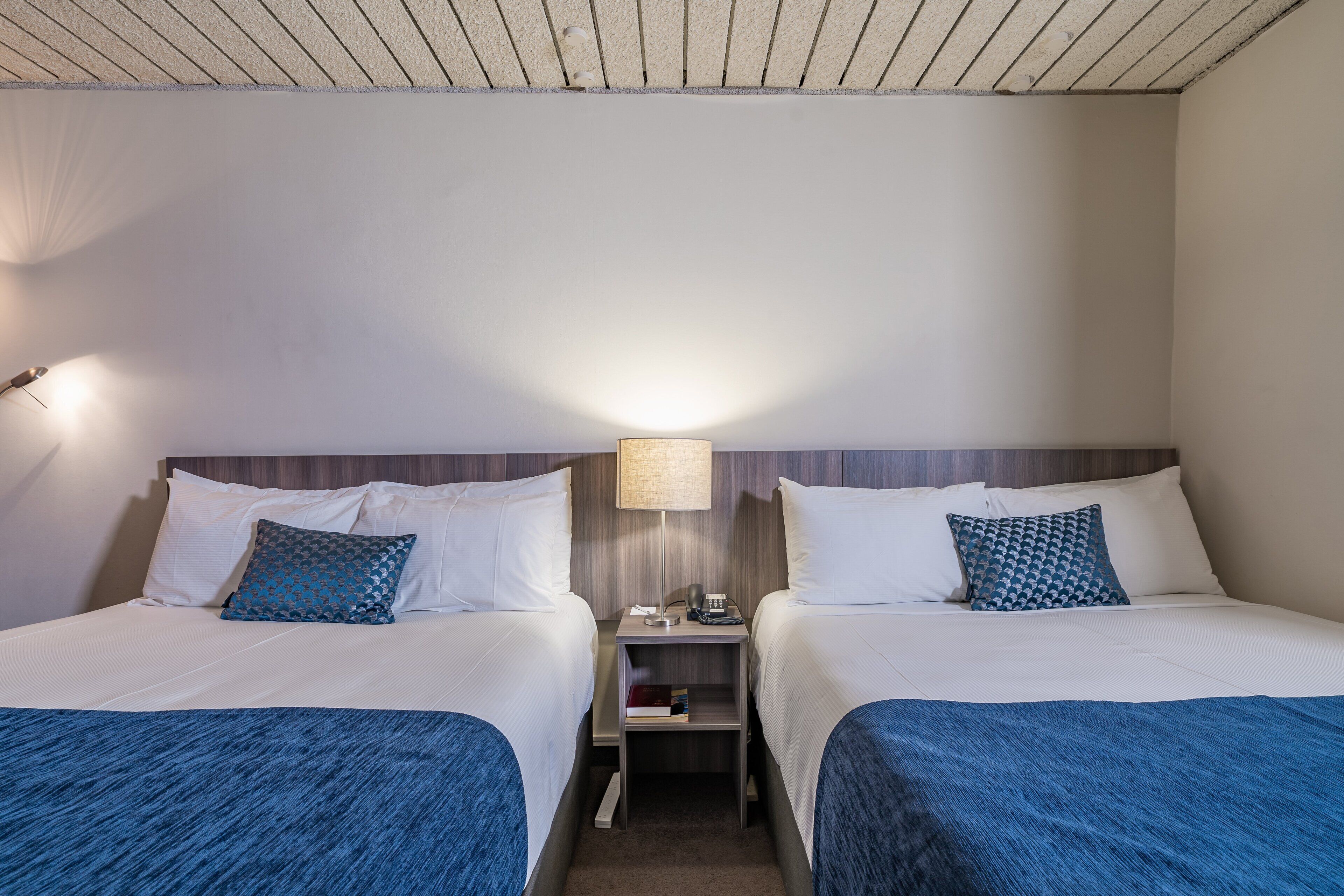 Una habitación de hotel con dos camas, una mesita de noche con lámpara y un pequeño estante.