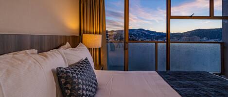 A hotel room with a large bed, a window with a view of a city and mountains, and a curtain.