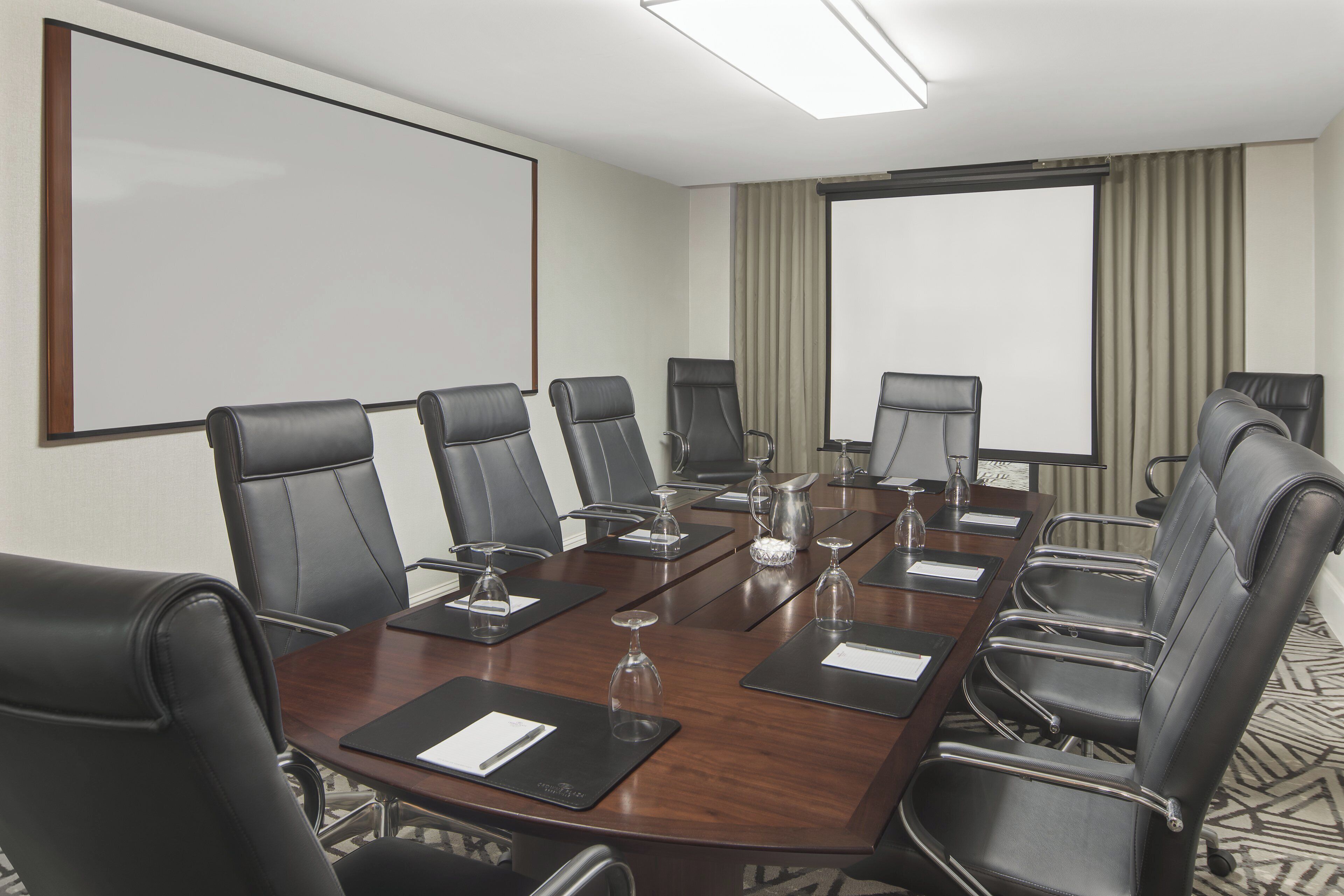 A conference room with a long table, multiple chairs, a projector screen, and a large window with curtains.