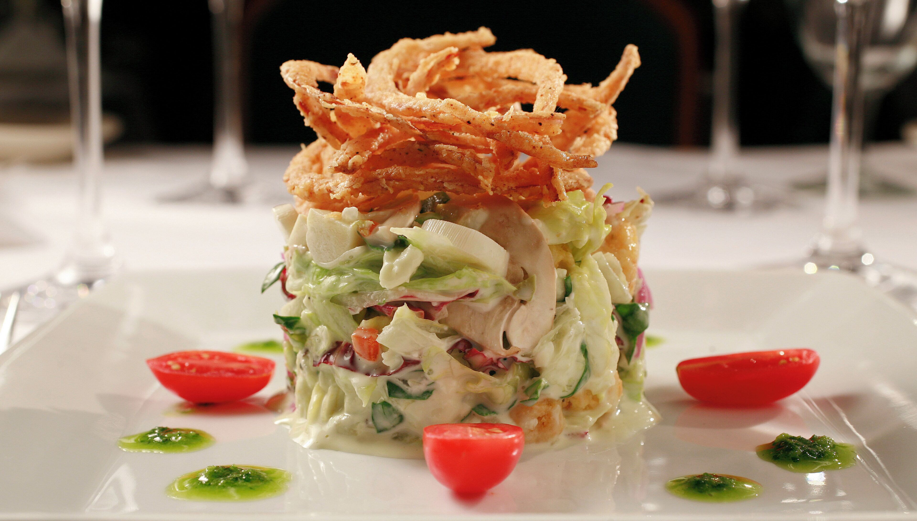A plated dish with a layered salad, garnished with red tomato slices and green sauce, served on a white plate.