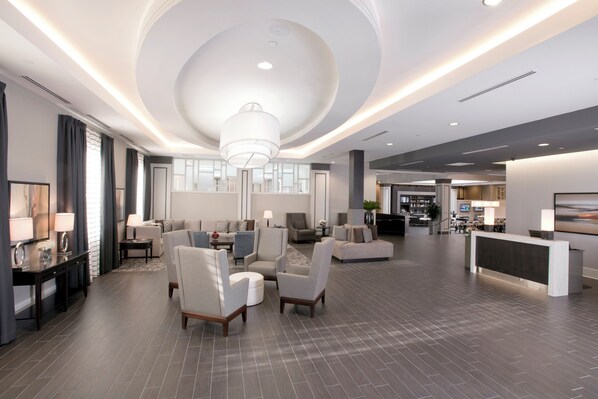 A spacious lobby with a modern design, featuring a reception desk, seating area with sofas and armchairs, and a large chandelier.