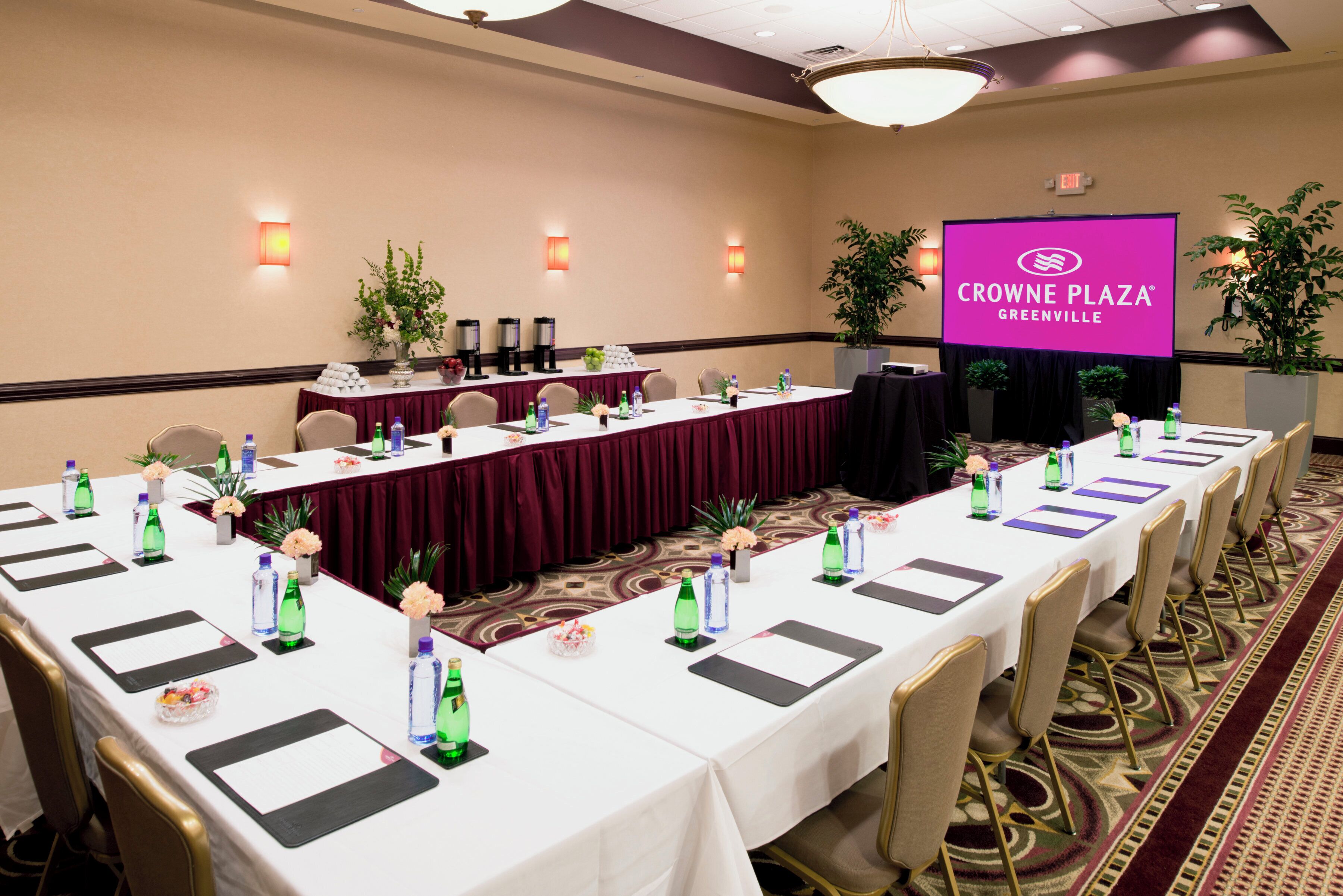 A conference room with a long table, chairs, water bottles, and a screen displaying the Crowne Plaza Greenville logo.