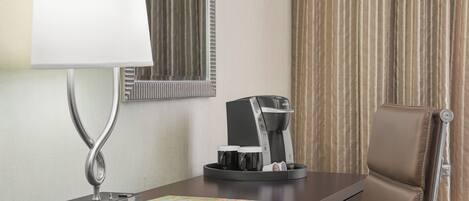 Une chambre d’hôtel équipée d’un bureau, d’une machine à café, d’une lampe, d’un miroir et d’une fenêtre avec des rideaux.