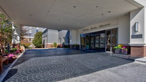 L’entrée d’un hôtel, agrémentée d’une enseigne accueillante, d’une allée couverte et d’un espace paysager avec des bancs et des fleurs.