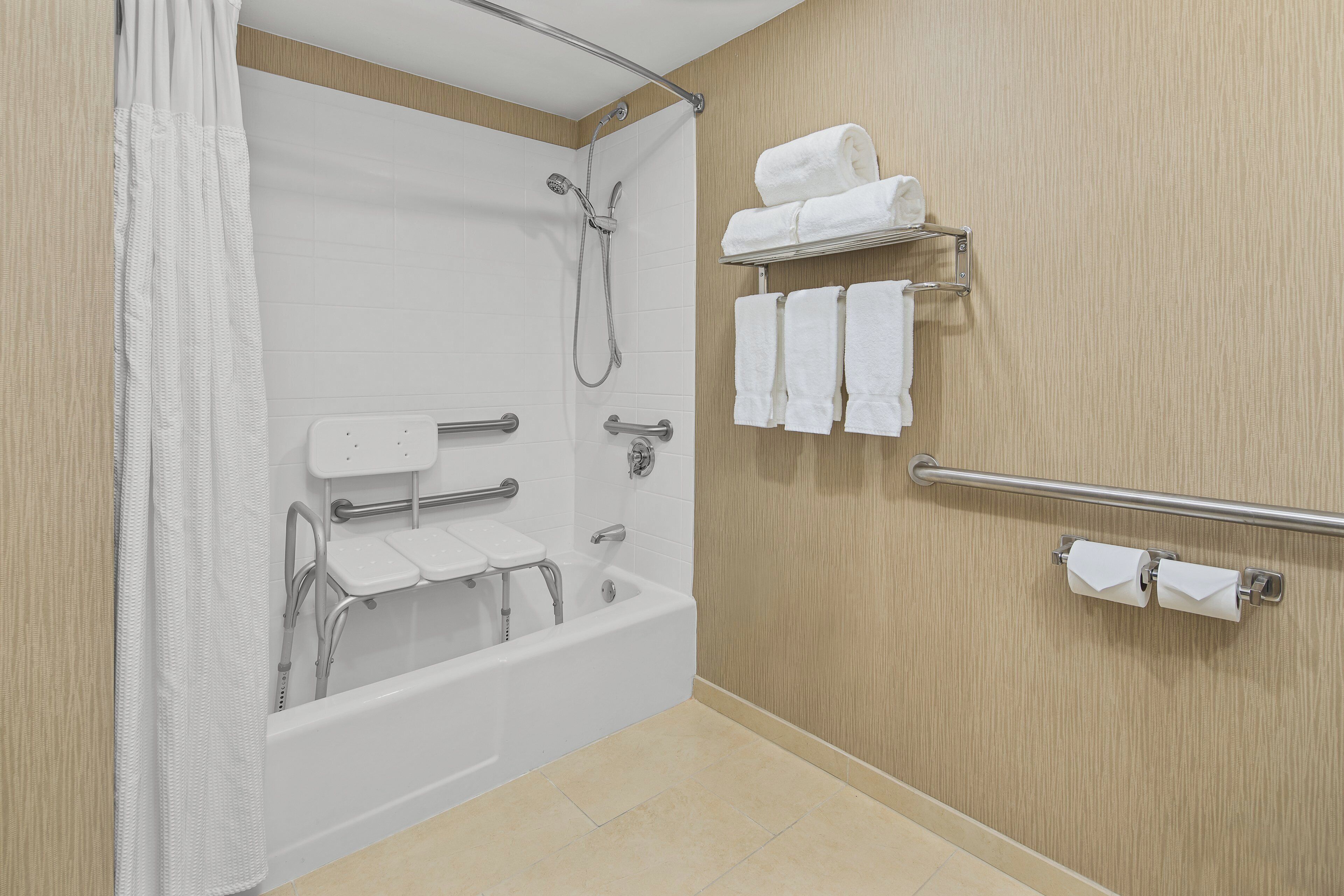 A bathroom with a bathtub, shower, grab bars, a toilet with a seat, and a towel rack.