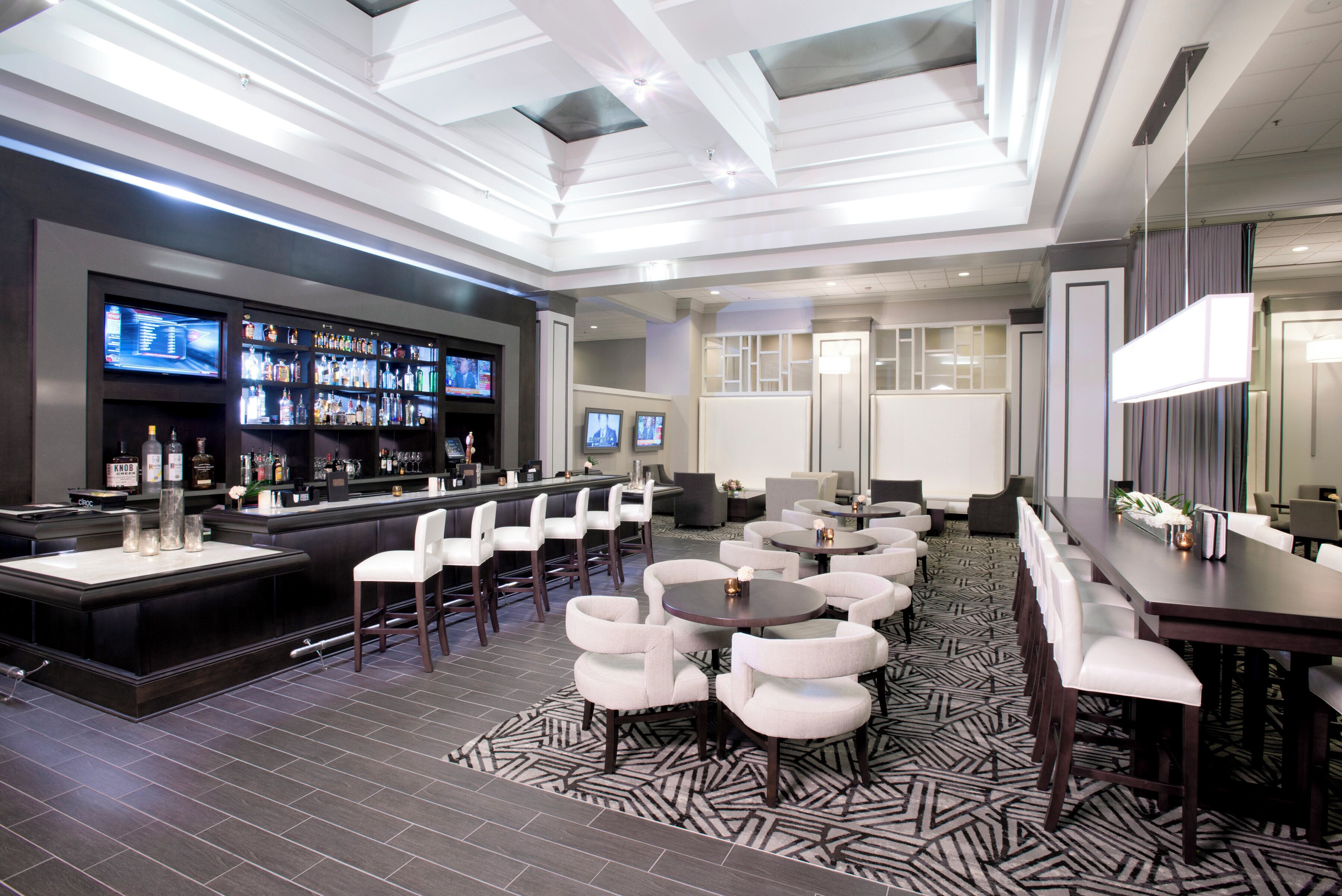 A modern bar area with a long counter, multiple screens, and a variety of liquor bottles.