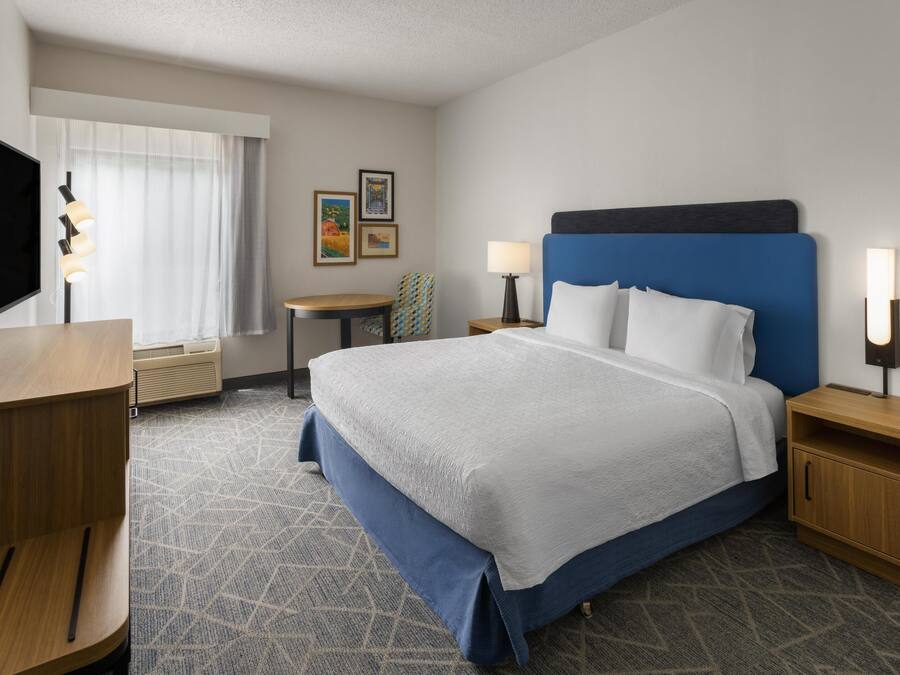 A hotel room with a bed, bedside tables, a desk, a chair, a TV, and artwork on the wall.