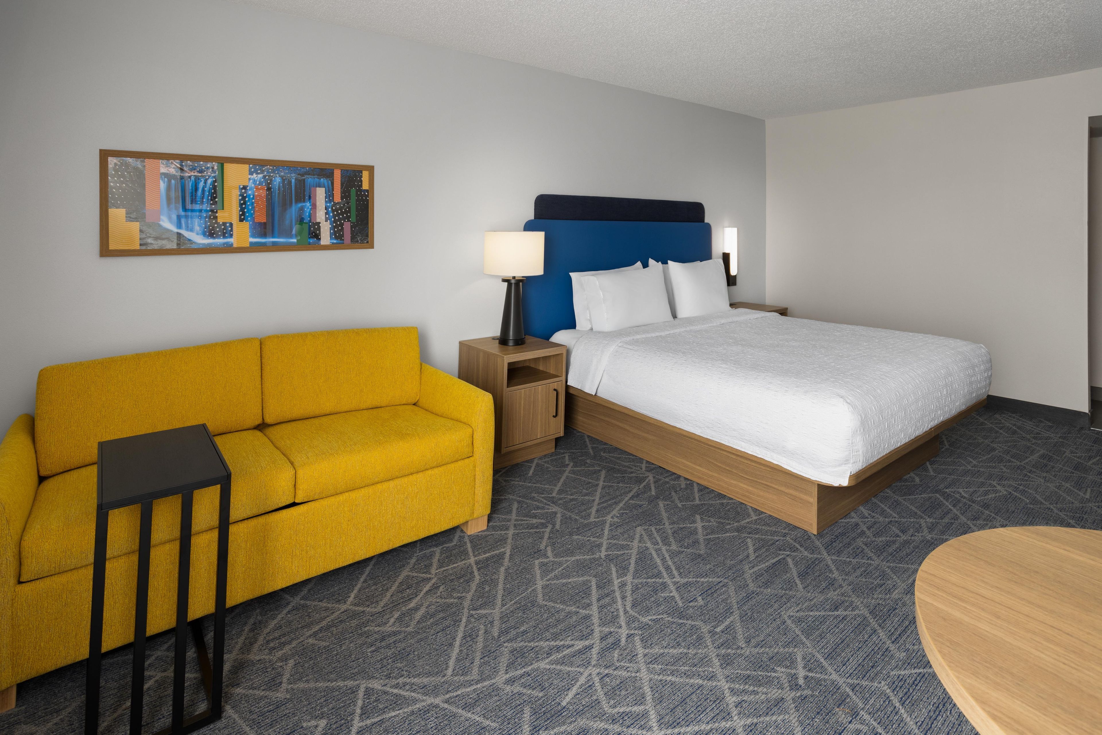 A hotel room with a bed, a sofa, a nightstand, a lamp, and a painting on the wall.