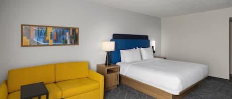 A hotel room with a bed, a sofa, a nightstand, a lamp, and a painting on the wall.