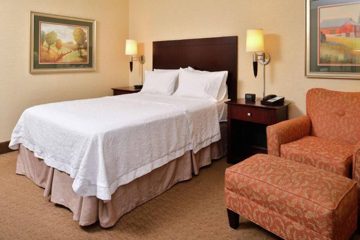Camera d'albergo con un letto grande, due comodini con lampade, una sedia e opere d'arte incorniciate alle pareti.