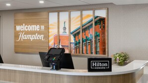 A hotel reception desk with a 'Welcome to Hampton' sign, a point-of-sale terminal, and a 'Hilton Honors' loyalty program sign. - Hampton Inn Staunton (Staunton)