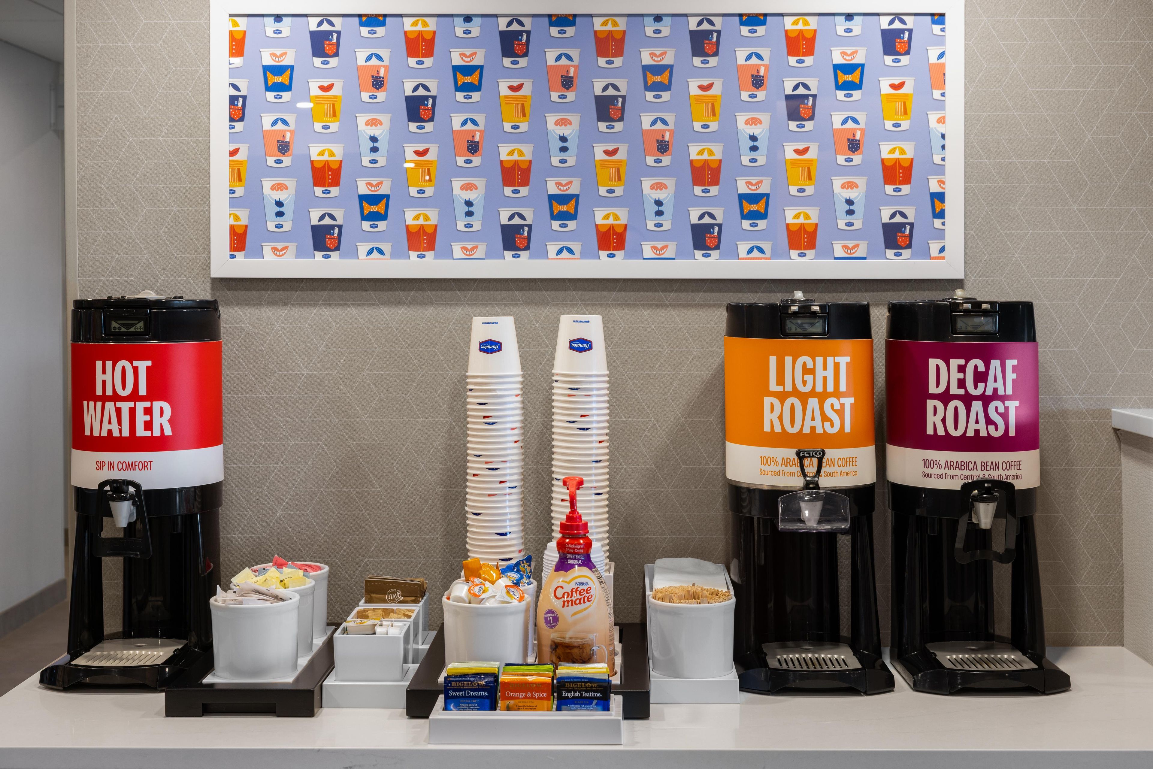 A coffee station with four dispensers labeled 'HOT WATER,' 'LIGHT ROAST,' and 'DECAF ROAST,' along with a variety of coffee pods and a wall display of coffee cup designs.